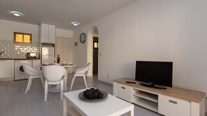 Un salon moderne avec un téléviseur, des meubles blancs et une cuisine en arrière-plan.