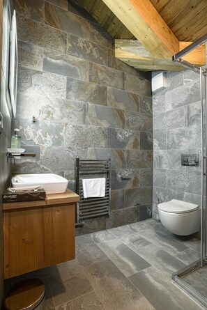 Un baño con pared de piedra, tocador de madera, lavamanos blanco, toallero y cabina de ducha con mampara de vidrio.