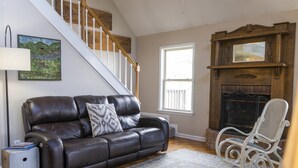 Un salon avec un canapé en cuir marron, un fauteuil à bascule blanc, une cheminée et un escalier.