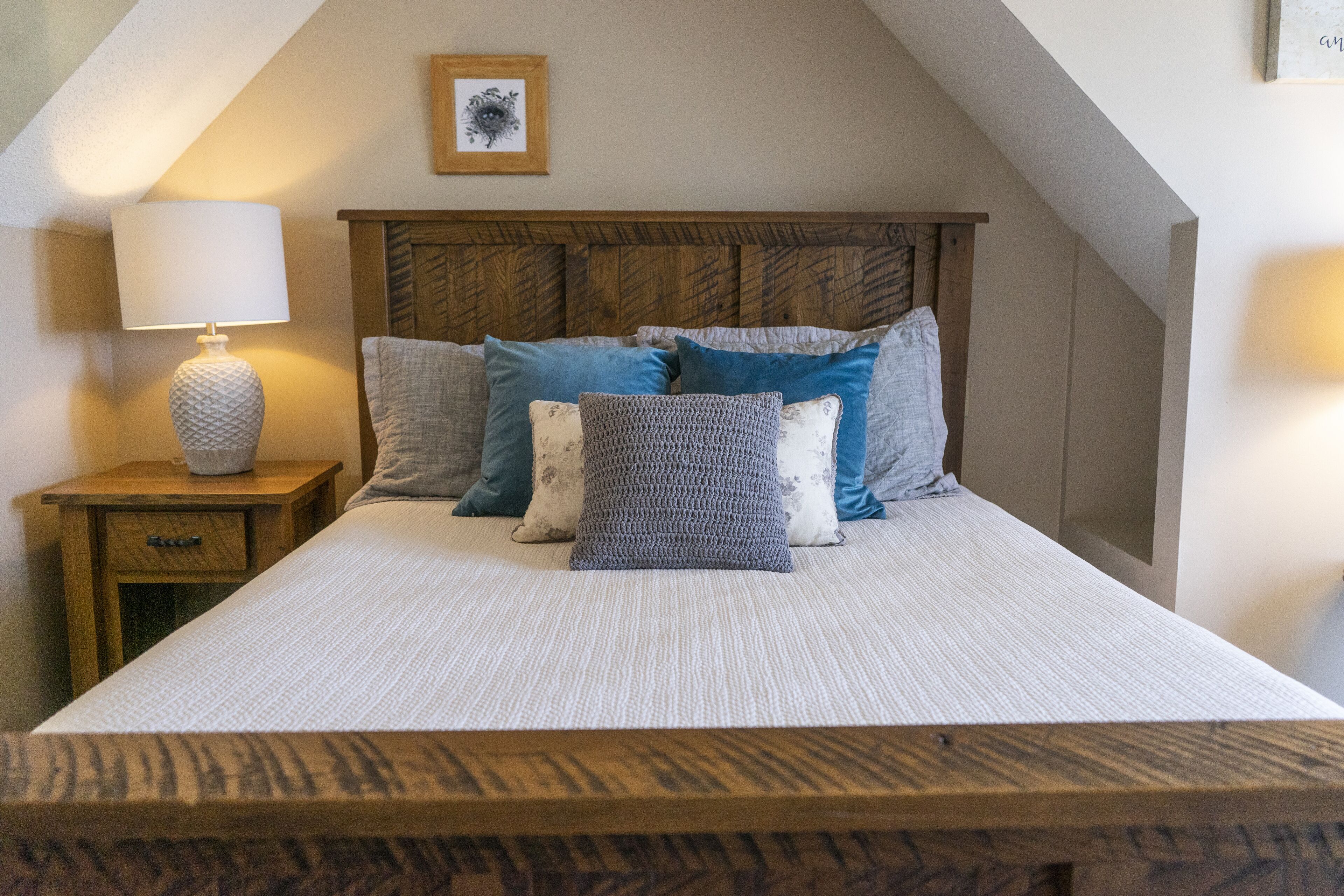 A bedroom with a wooden headboard, a bedside table with a lamp, and a bed with multiple pillows.