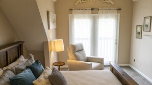 Une chambre à coucher avec un grand lit, une chaise, une lampe et une vue sur l’extérieur par une fenêtre avec des rideaux.