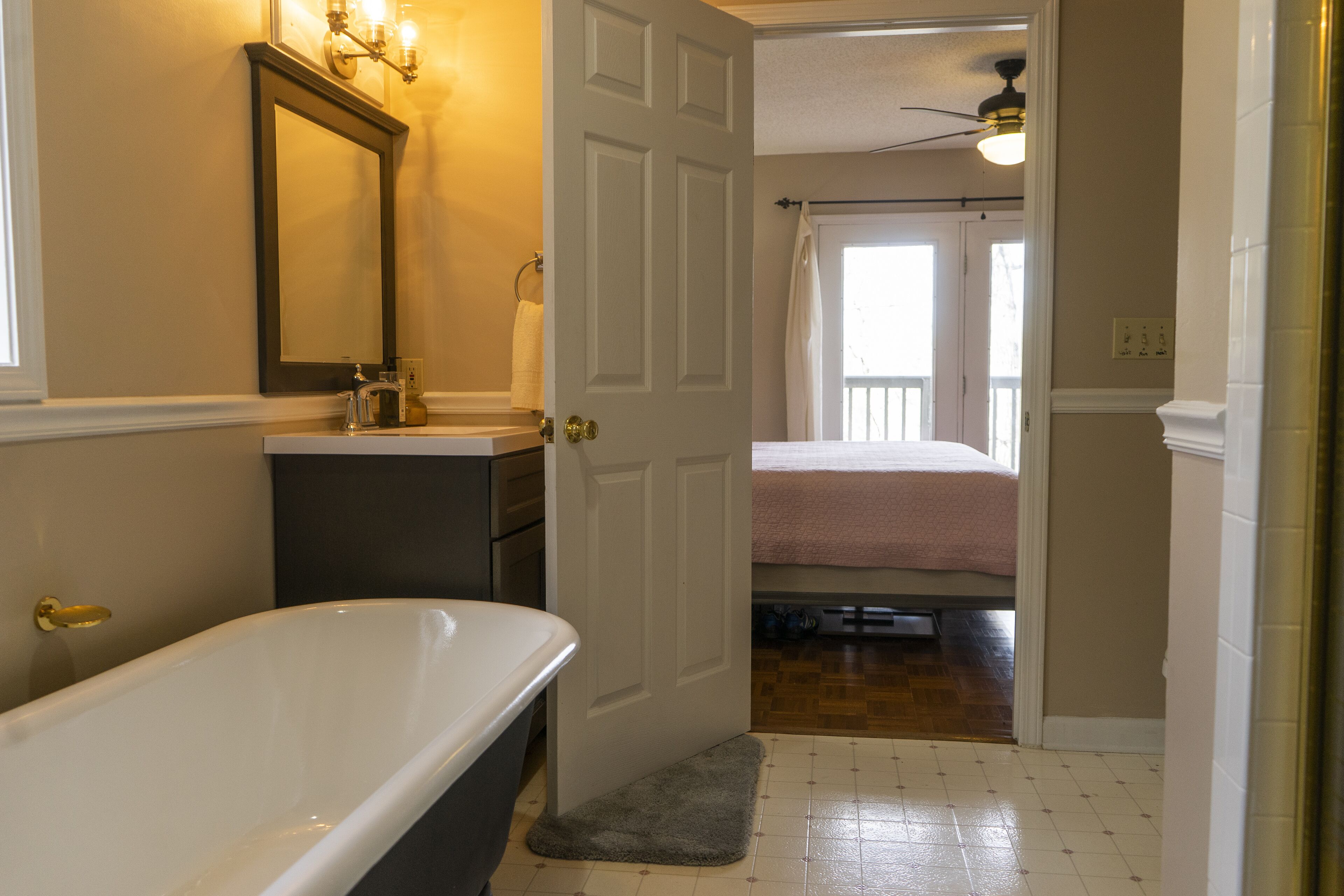 A bathroom with a bathtub, a sink, and a mirror. There is a door leading to a bedroom with a bed and a window.