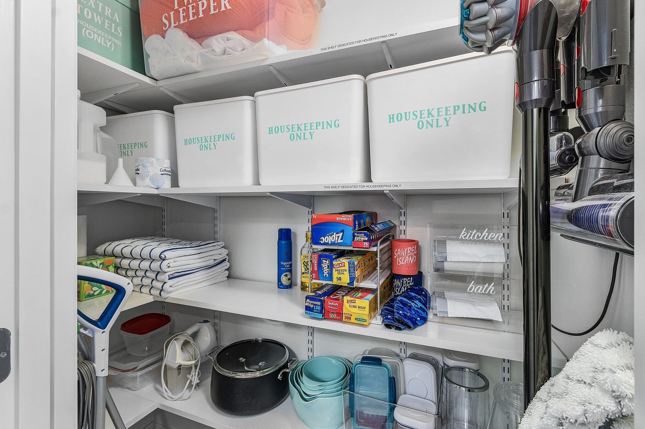 A neatly organized storage area with labeled bins for housekeeping items, kitchen and bath supplies, and neatly folded towels.
