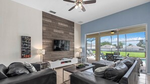 A modern living room with a flat-screen TV, leather sofas, a coffee table, and a large window offering a view of a pool and outdoor furniture.