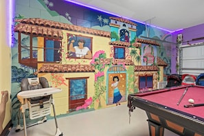 Une pièce équipée d’une table de billard, d’une chaise haute et ornée d’une fresque représentant une scène colorée avec des bâtiments et des personnages.