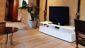 Una sala de estar con piso de madera, un mueble blanco para entretenimiento, un televisor de pantalla plana grande, una planta en maceta y una silla de madera.