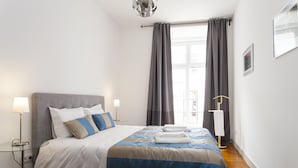 Una cama bien hecha con colcha azul y blanca, dos lámparas de noche y una ventana con cortinas.