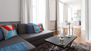 Una sala de estar moderna con un sofá gris, cojines de colores, una mesa de centro y vista hacia la zona de la cocina.