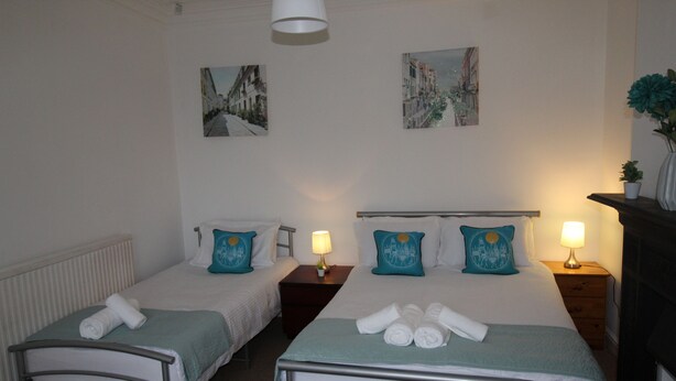 A hotel room with two single beds, each with a pillow and a bedside table with a lamp. There are two paintings on the wall, a radiator under the beds, and a vase with a plant on a dresser. - Stafford House (Sheffield)