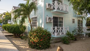 A two-story house with a balcony, air conditioning units, and a sandy front yard with plants.