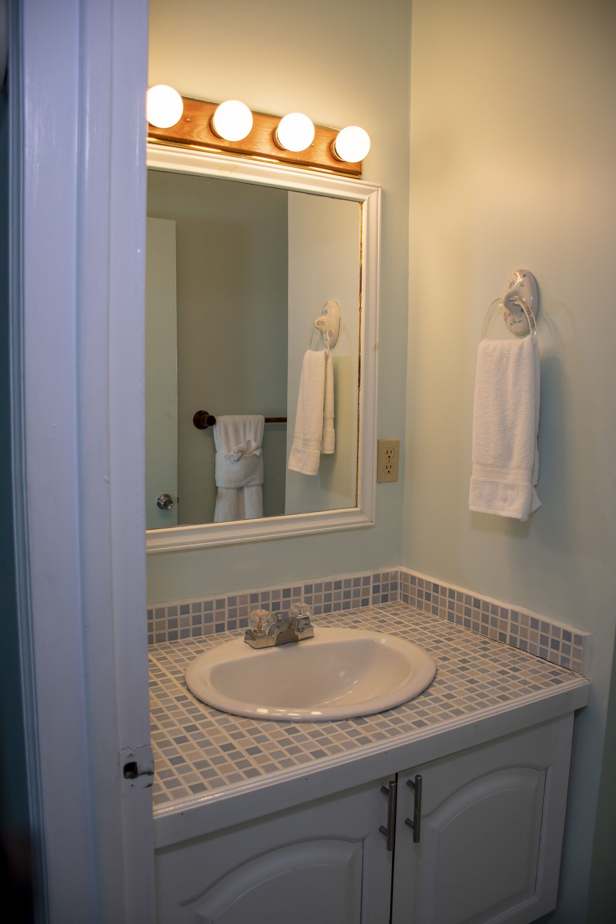 Un baño con un lavamanos blanco, un espejo con luces y dos toallas colgadas en ganchos.