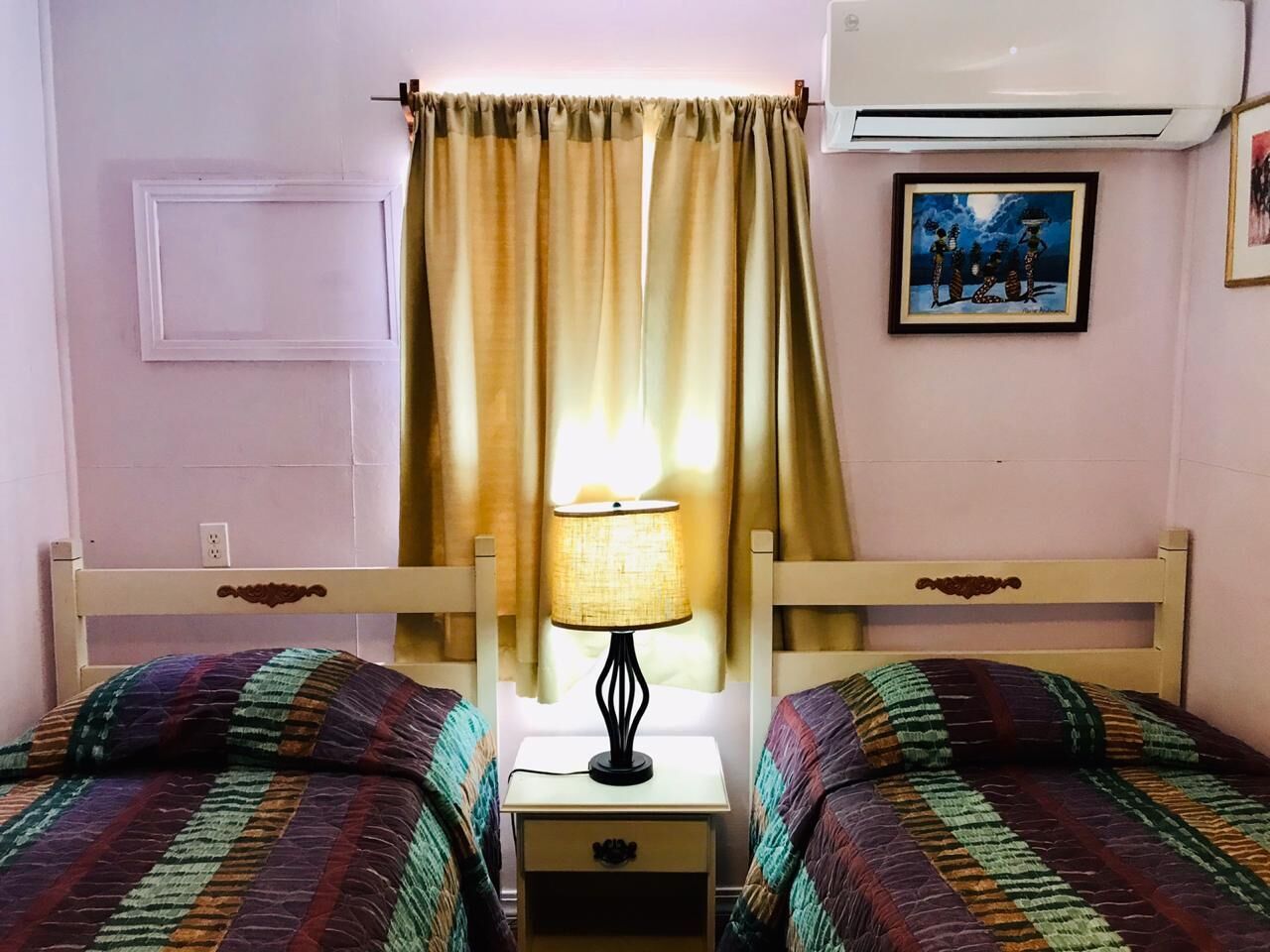 Een kamer met twee bedden, een nachtkastje met lamp, een raam met gordijnen, een airconditioner en een ingelijst schilderij aan de muur.
