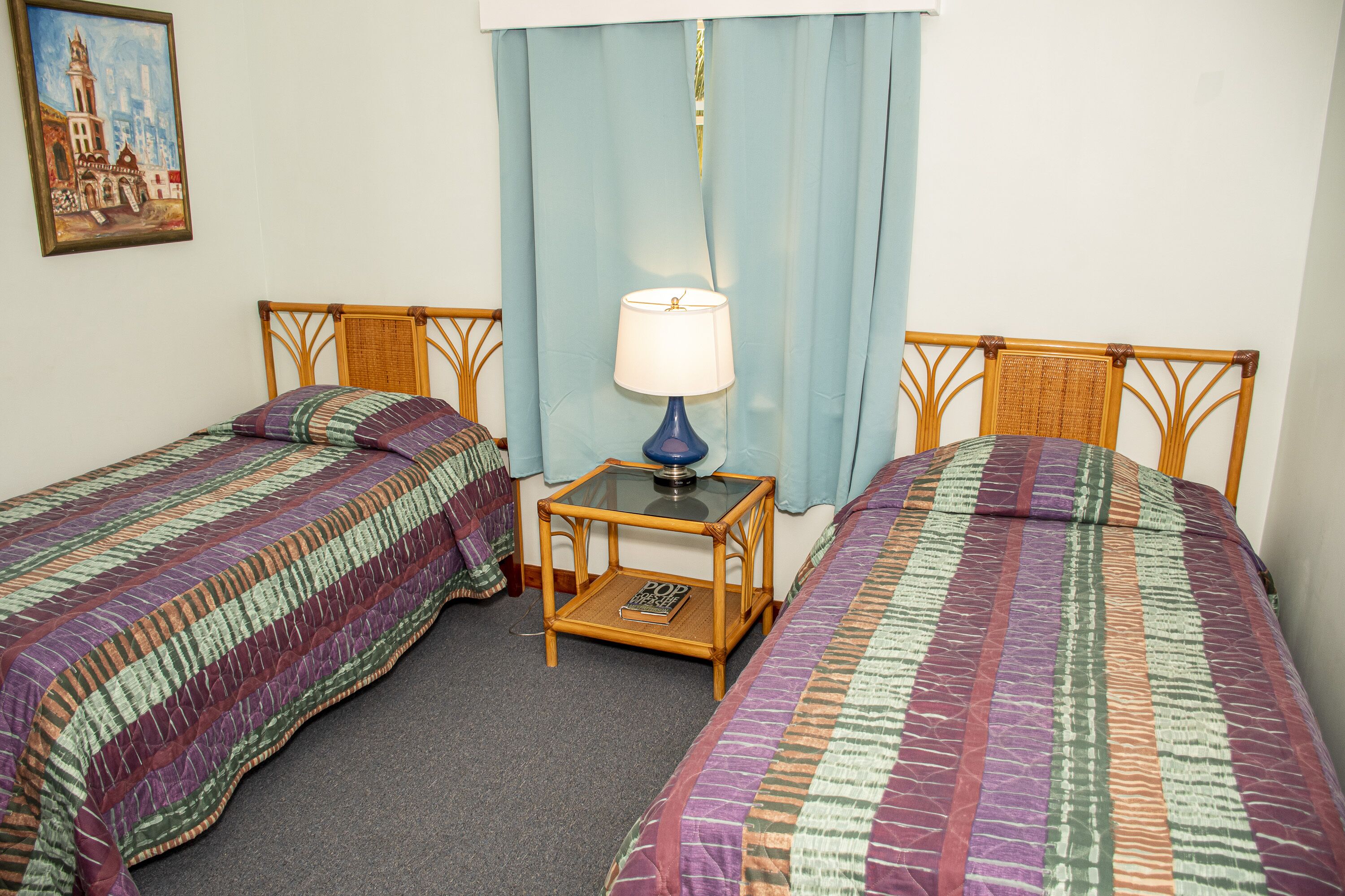 A room with two single beds, a small table with a lamp, and a painting on the wall.