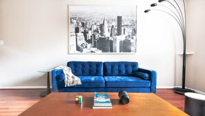 A living room with a blue sofa, a coffee table with a book and a Rubik's cube, and a cityscape painting on the wall. - COMFY COZY MED CENTER HOME (Houston)