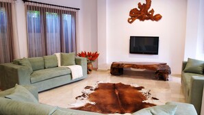 Uma sala de estar com um sofá verde, uma mesa de centro de madeira, um tapete de couro e uma televisão de tela plana na parede.