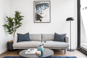 Un salon moderne avec un canapé gris, des coussins à rayures, un lampadaire noir, une grande plante en pot et une œuvre d’art encadrée accrochée au mur.