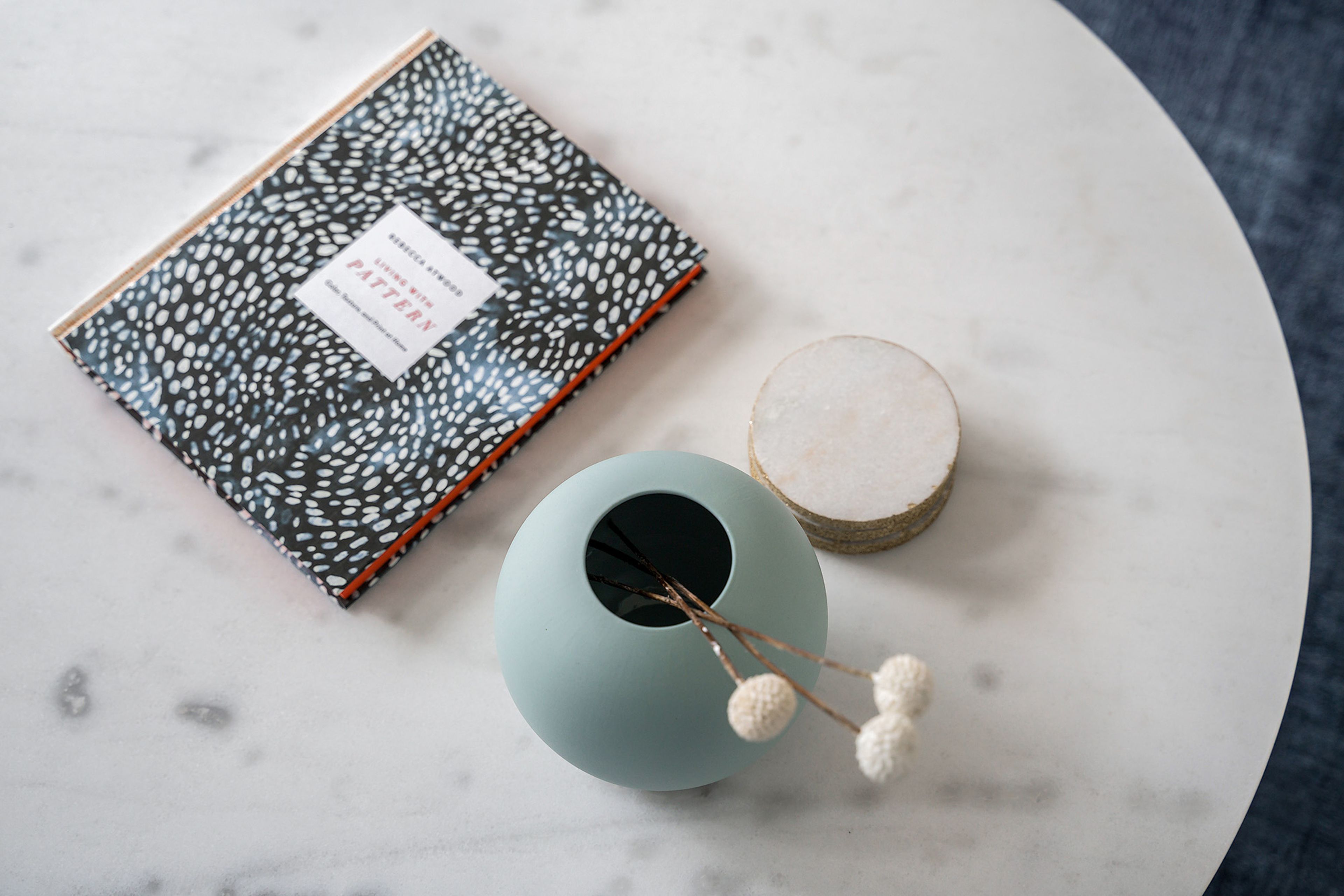 A book with a patterned cover, a round table with a vase and a small round object on it.
