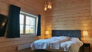 Et soveværelse i en træhytte med en seng, to sengeborde med lamper, et vindue med gardiner og en hængende lampe.