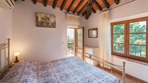 A bedroom with a wooden ceiling, a bed with a floral quilt, a painting on the wall, and a window with a view of greenery.