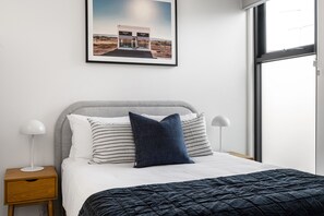 Ein ordentlich bezogenes Bett mit einer dunklen Steudecke, zwei Nachttischlampen, einem Nachttisch und einem gerahmten Bild von einem Prada-Geschäft an der Wand.