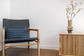 A wooden chair with a dark cushion and a striped pillow, positioned next to a wooden side table with a vase of dried branches. - Axel Apartments 203 The Bonfield (Glen Iris)