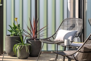 A patio area with a wicker chair, a potted plant, and a table with a mug and a book. - Axel Apartments 203 The Bonfield (Glen Iris)