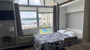 A hotel room with a bed, a desk, and a large window offering a view of a body of water and buildings. - Lakefront Condo on Crooked Lake, Convenient Location to Northern Michigan Cities (Conway)
