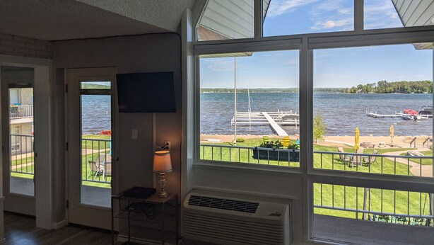 A room with a large window overlooking a lake, a TV mounted on the wall, and a table with chairs. - Lakefront Condo on Crooked Lake, Convenient Location to Northern Michigan Cities (Conway)