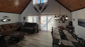 A cozy living room with a high ceiling, wooden beams, a fireplace, a dining area with a table set for four, and a large window with blinds. - Lakefront Chalet - Badwater Lodge - Gorgeous Lake Views (Florence)