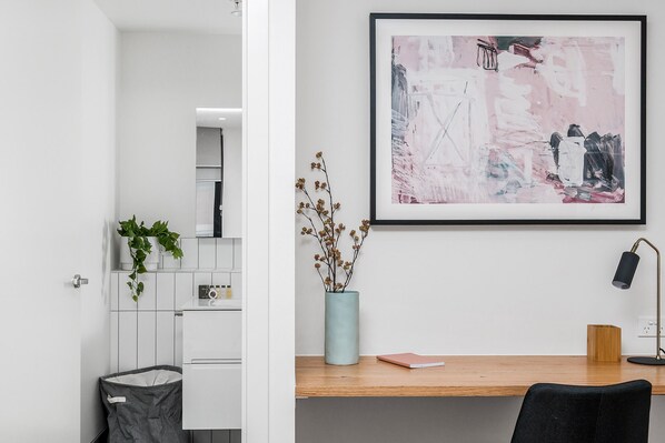 A modern interior with a desk, a framed abstract painting, a vase with dried flowers, and a trash bin.