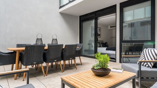 A modern outdoor seating area with wicker chairs and a wooden table, a potted plant, and a view into a bedroom through a glass door.