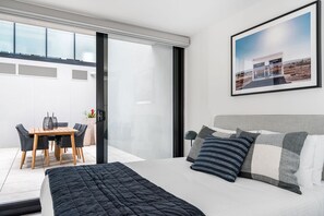 A modern bedroom with a large bed, a dining area with chairs, and a view through a sliding glass door. - Axel Apartments G02 The Faircroft (Glen Iris)
