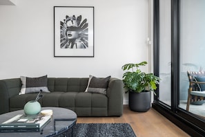 A modern living room with a grey sofa, a coffee table, a framed artwork, a potted plant, and a view through a large window. - Axel Apartments G02 The Faircroft (Glen Iris)