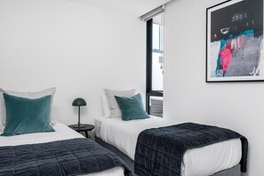 A bedroom with two beds, a nightstand, a lamp, and a window with blinds. - Axel Apartments G02 The Faircroft (Glen Iris)