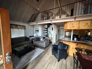 A spacious living area with a sofa, coffee table, and a dining table with chairs. A spiral staircase leads to an upper level. - Dog Friendly Cute cabin on quiet lot with lots of pines. (Munds Park)