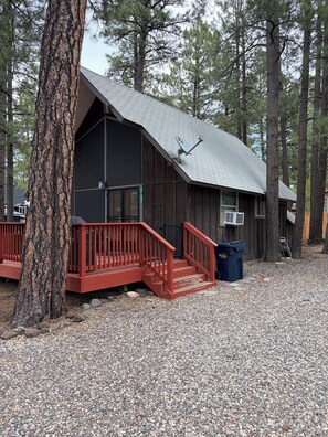 Une cabane avec un escalier rouge, une antenne parabolique et une allée en gravier, le tout entouré d’arbres.