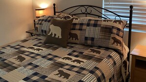 A bedroom with a bed covered in a checkered quilt, a headboard, a bedside table, and a window with blinds.