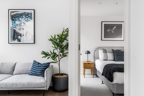 A modern bedroom with a grey sofa, a potted plant, a bedside table with a lamp, and a bed with a grey headboard. - Axel Apartments 103 The Hadley (Glen Iris)