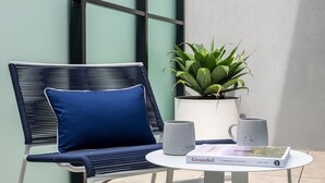 Ein Stuhl mit blauer Sitzkissen, ein Blumentopf, zwei Tassen und ein Buch mit dem Titel 'Grounded' auf einem kleinen Tisch.