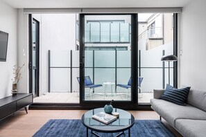 A modern living room with a sofa, a coffee table with a book, and a view through a glass door to an outdoor seating area. - Axel Apartments 103 The Hadley (Glen Iris)
