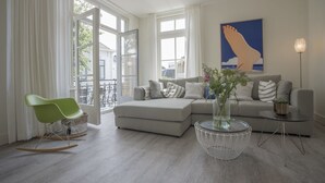 Un salon moderne avec un canapé gris, un fauteuil à bascule vert, une table basse en verre et une grande fenêtre ornée de rideaux blancs.