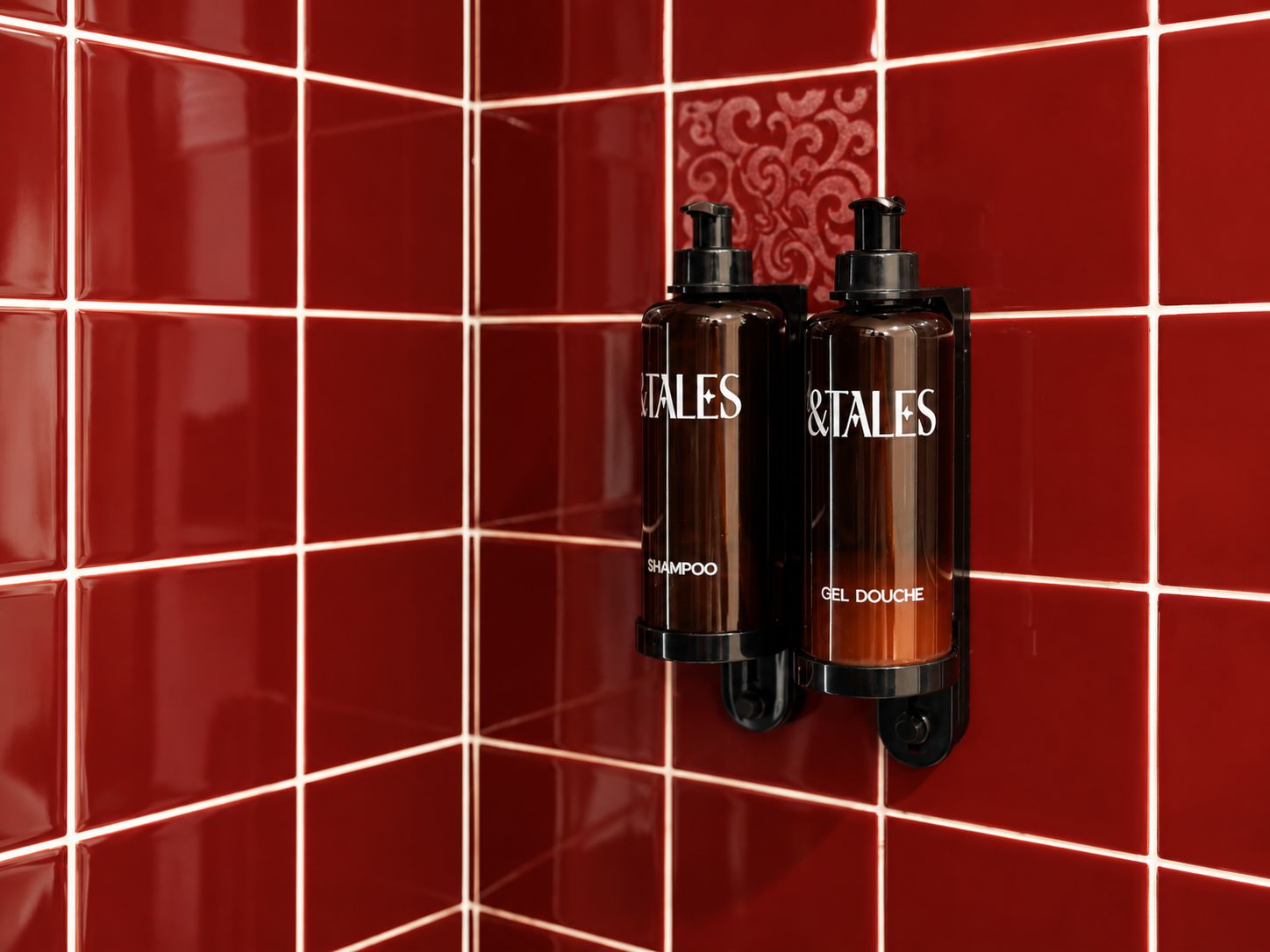 Dos dispensadores de artículos de tocador con la etiqueta 'Tales', uno con la leyenda 'Champú' y otro con 'Gel de ducha', sobre una pared de baldosas rojas.