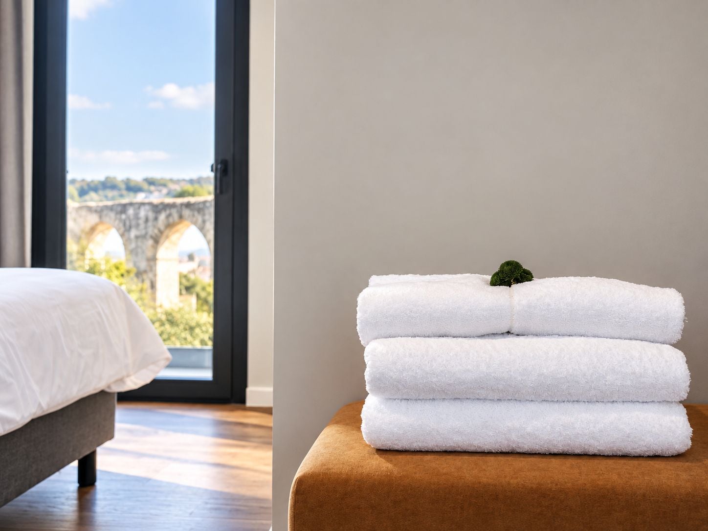 Una camera d'albergo con un letto, una finestra vista su un ponte di pietra e una pila di asciugamani bianchi su un pouf marrone.