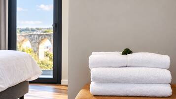 A hotel room with a bed, a window with a view of a stone bridge, and a stack of white towels on a brown ottoman.