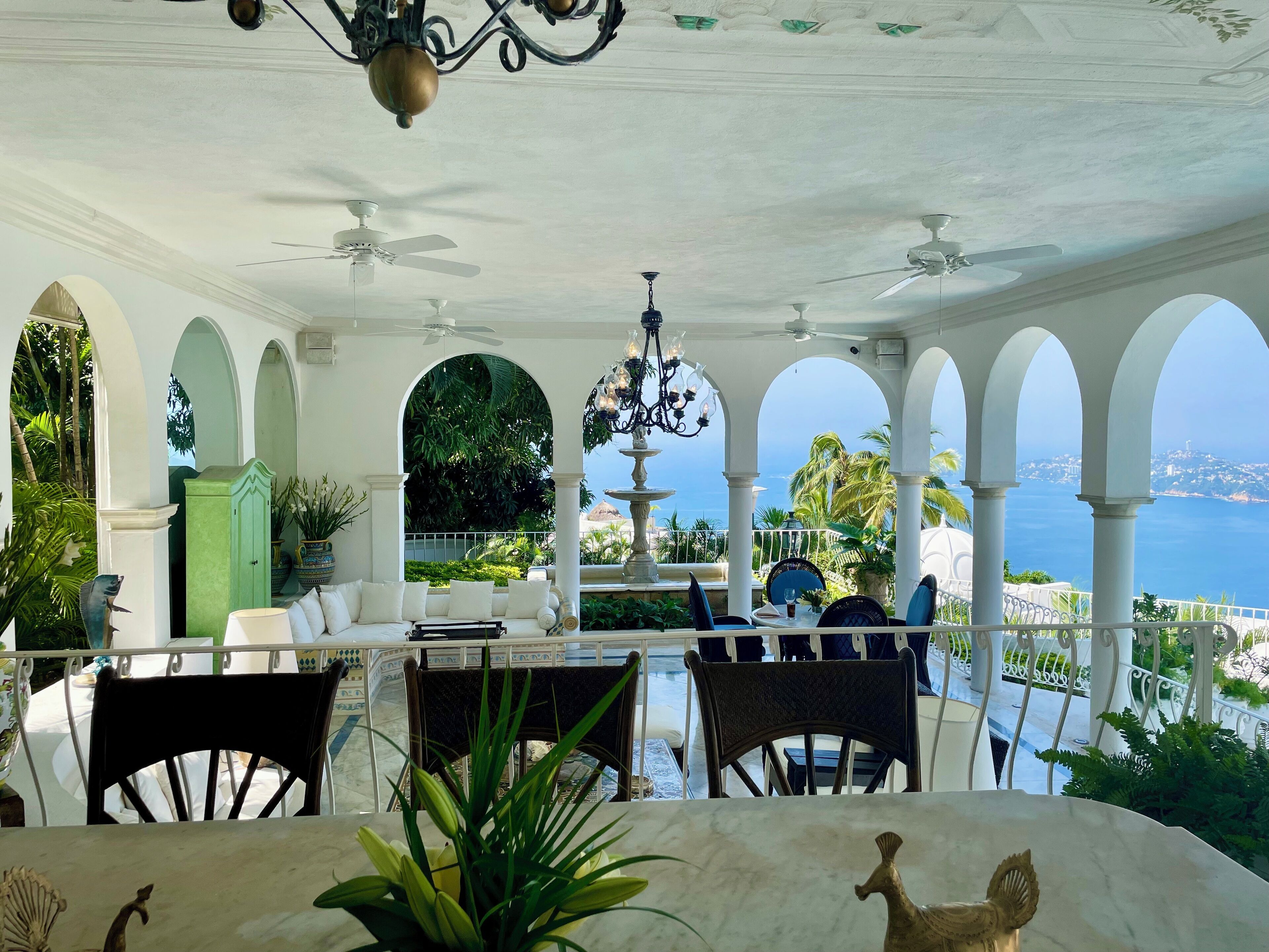Un espacio de estar al aire libre cubierto con columnas blancas, ventiladores de techo y vista al mar.