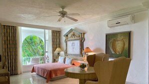 Une chambre d’hôtel comprenant un lit, une table ronde, une chaise, un ventilateur de plafond, un appareil de climatisation et un balcon avec vue sur la verdure.