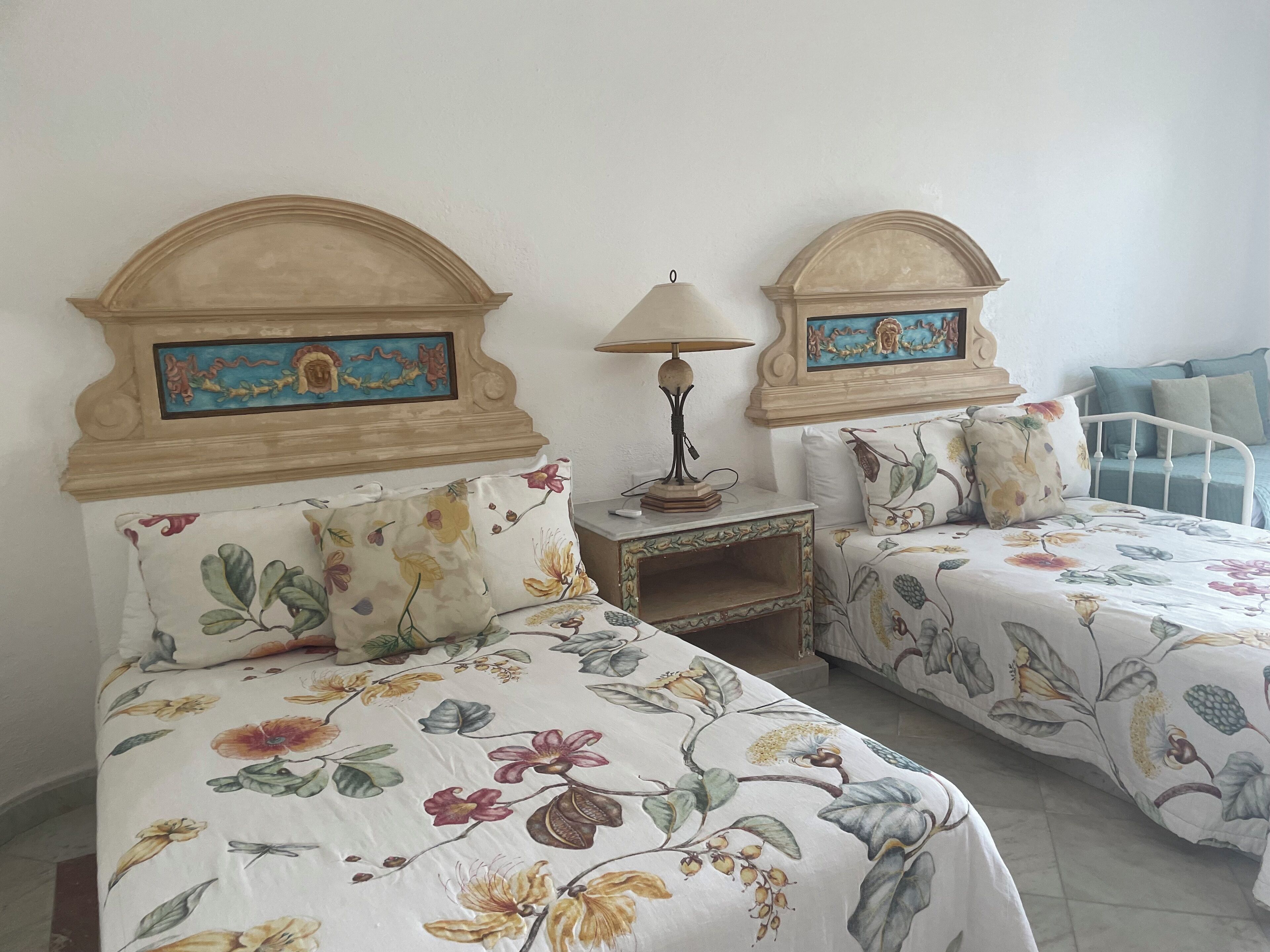 A room with two beds, each with floral bedding and headboards, a nightstand with a lamp, and a painting above the headboards.