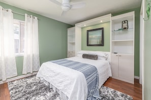 Une chambre à coucher comprenant un lit, un ventilateur de plafond, une fenêtre avec des rideaux et des étagères intégrées.