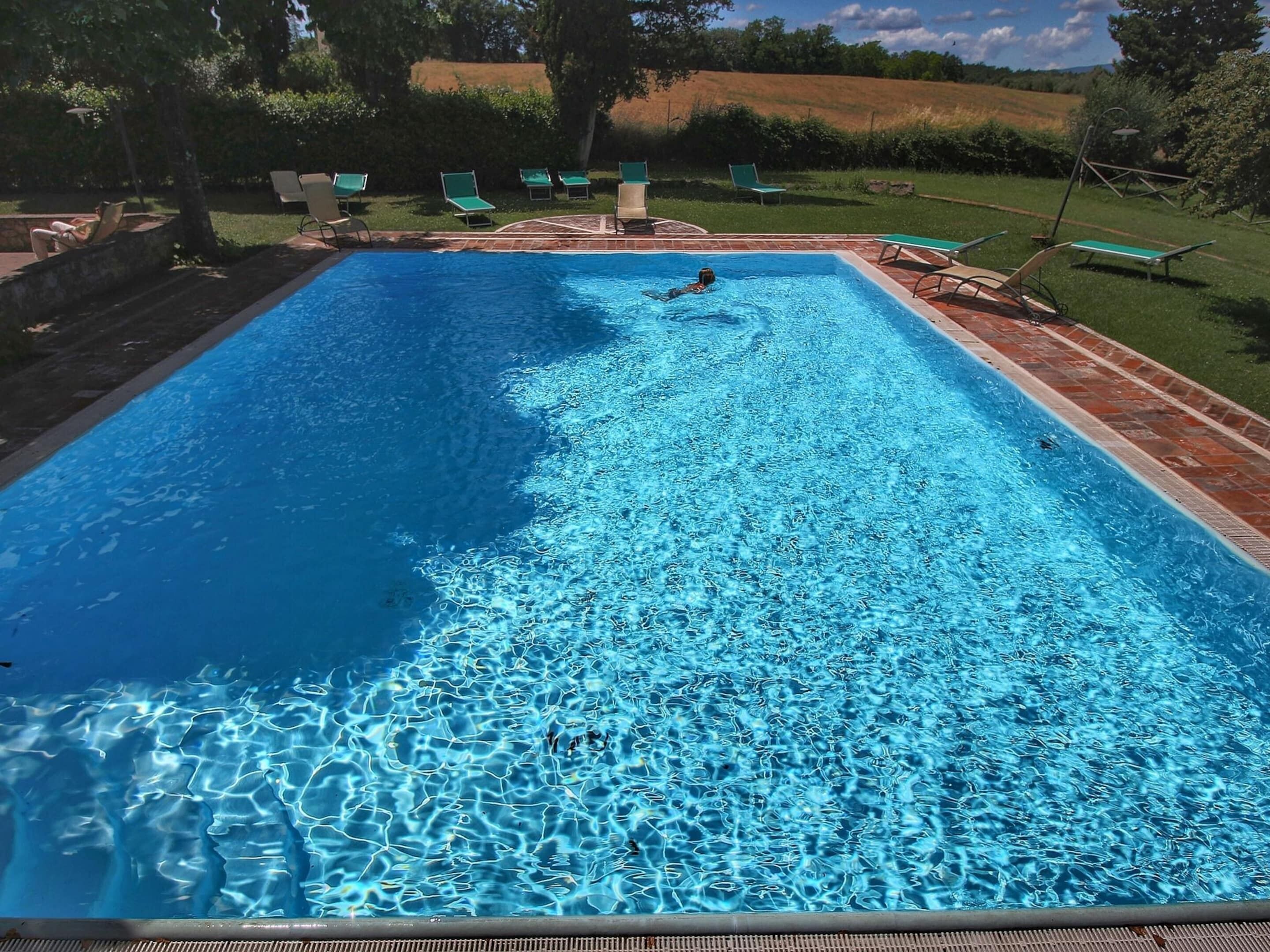 A rectangular swimming pool with clear blue water, surrounded by a brick deck and green lounge chairs.
