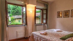 A bedroom with a wooden floor, a bed with floral bedding, a nightstand with a candle, and a view of greenery through the window. - Holiday Home for 8 Guests (Siena)